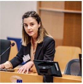 A woman with brown hair, wearing a dark blazer, sits at a desk with a microphone, computer, and name badge in a conference room setting, appearing to speak or present.