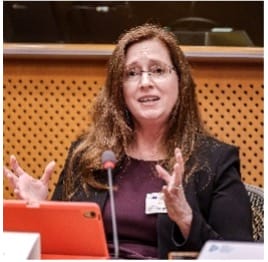 A woman with long brown hair and glasses speaks while gesturing with her hands at a conference table. She is wearing a black blazer and sits in front of a microphone and an orange tablet.