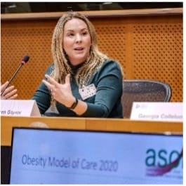 A woman with long blonde hair speaks whilst seated at a conference table. In the foreground, a screen displays the text “Obesity Model of Care 2020” and part of the ASO logo.