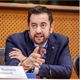 A man in a suit is sitting at a table, speaking and gesturing with his right hand. There is a microphone and a nameplate partially visible in front of him, and a wooden panel with holes is in the background.