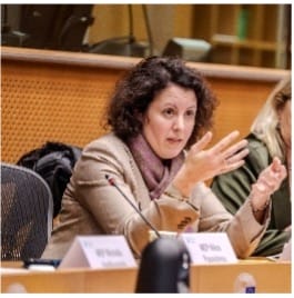 A woman with curly dark hair, wearing a beige blazer and a pink scarf, speaks while gesturing with her hand at a conference table in a meeting room. Another person sits beside her. Nameplates are visible on the table.