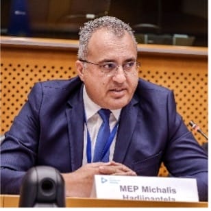 A man in a suit and tie sits at a conference table with a microphone and nameplate reading "MEP Michalis Hadjipantela." A perforated wooden panel is visible in the background.