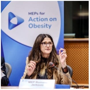 A woman with long brown hair and glasses speaks whilst seated at a table in front of a banner that reads "MEPs for Action on Obesity." She wears a tan jacket and gestures with her hands.