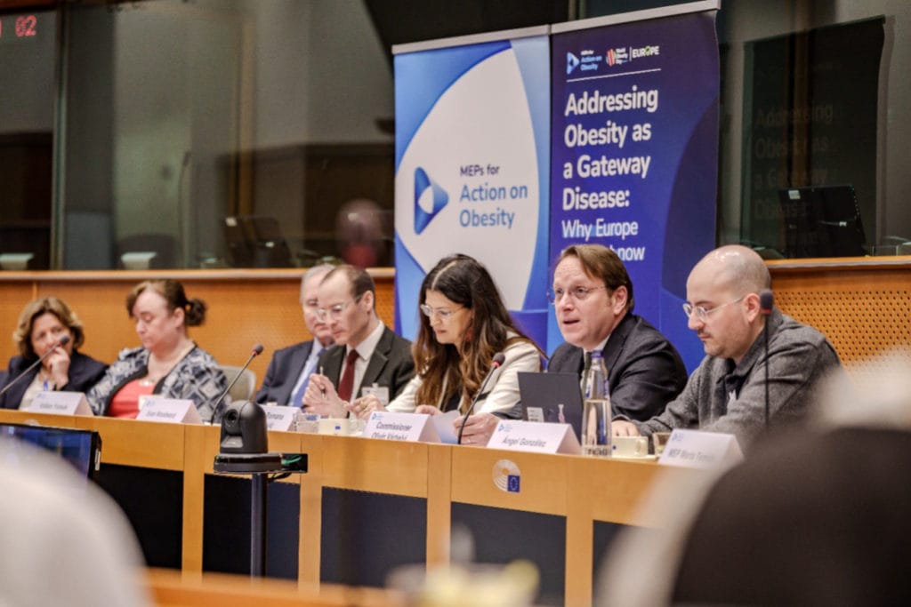 Six people sit at a panel table in a conference room. A banner behind them reads “Addressing Obesity as a Gateway Disease: Why Europe Needs to Act Now.” Microphones, nameplates, and water bottles are on the table.